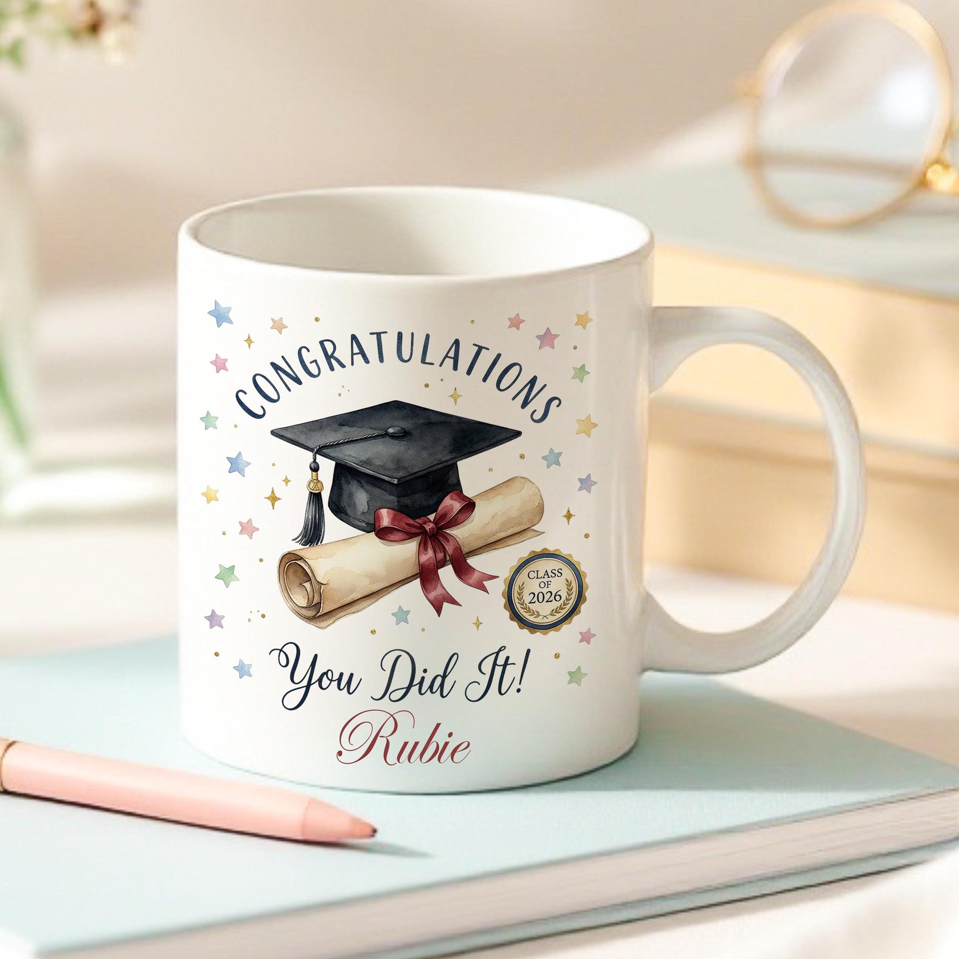 White ceramic mug printed with 'Congratulations You Did It!' in script, a watercolour graduation cap and diploma scroll with a Class of 2026 seal, pastel confetti stars, and the personalised name 'Rubie' in red script