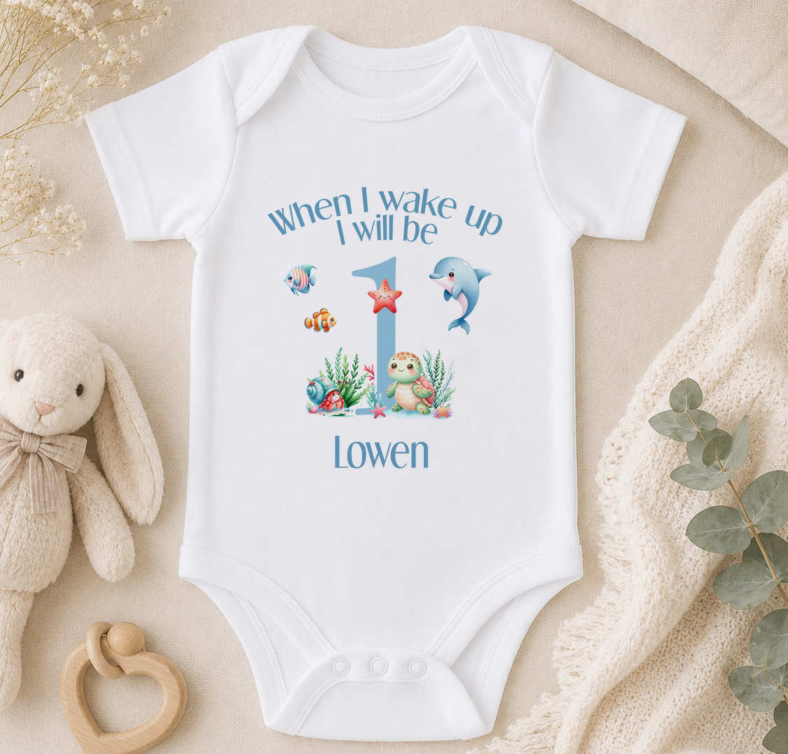 A white onesie with a personalized message and illustrations of a bunny, fish, and turtle, along with the text "When I wake up I will be 1 Lowen".