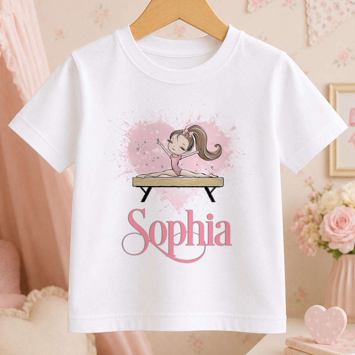 A white t-shirt with a graphic design featuring a young girl on a balance beam, surrounded by pink and white splatter effects. The word "Sophia" is written below the graphic.