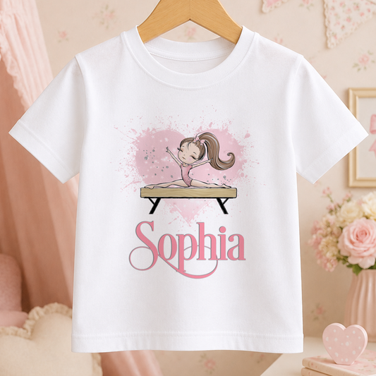 A white t-shirt with a graphic design featuring a young girl on a balance beam, surrounded by pink and white splatter effects. The word "Sophia" is written below the graphic.
