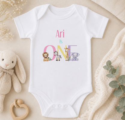 A white onesie with the text "Ari is ONE" printed on it, surrounded by various stuffed animals and a beige blanket.
