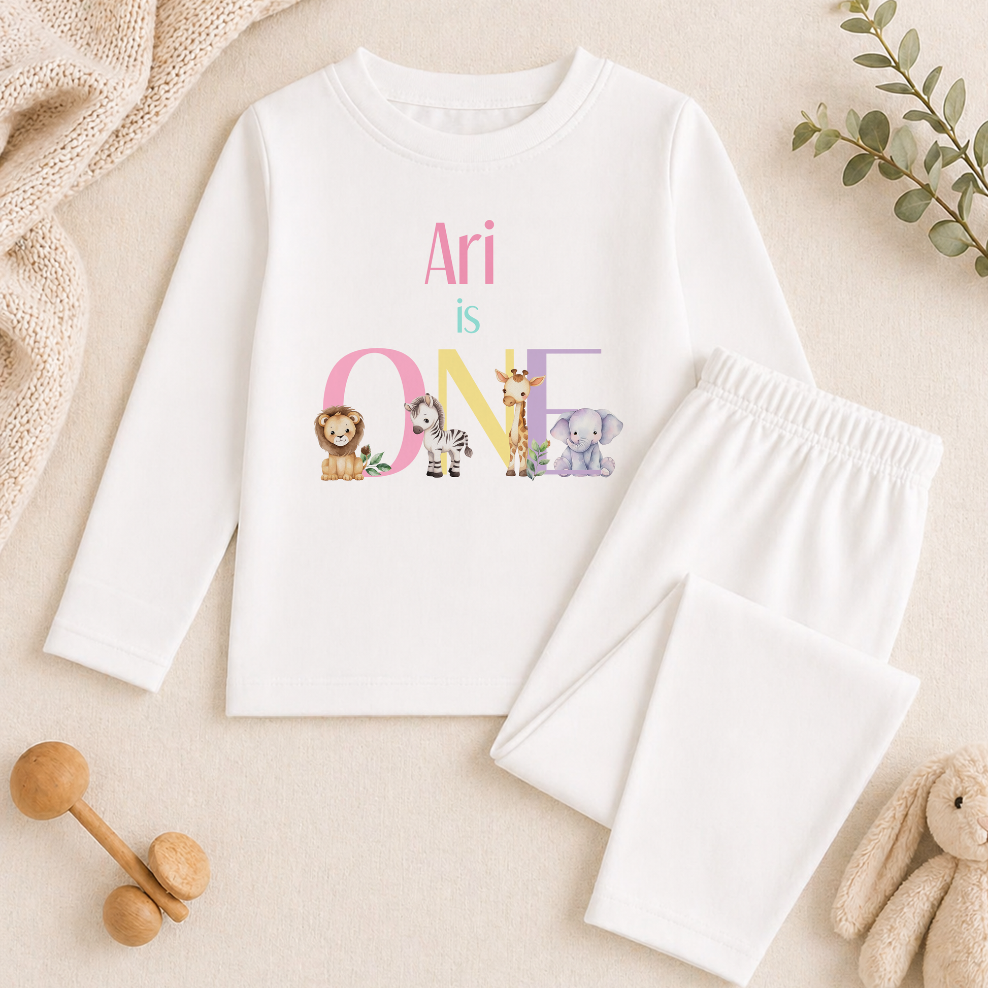 A white long-sleeved shirt with a personalized text and illustrations of various animals, along with a pair of white pants, are displayed on a beige surface.