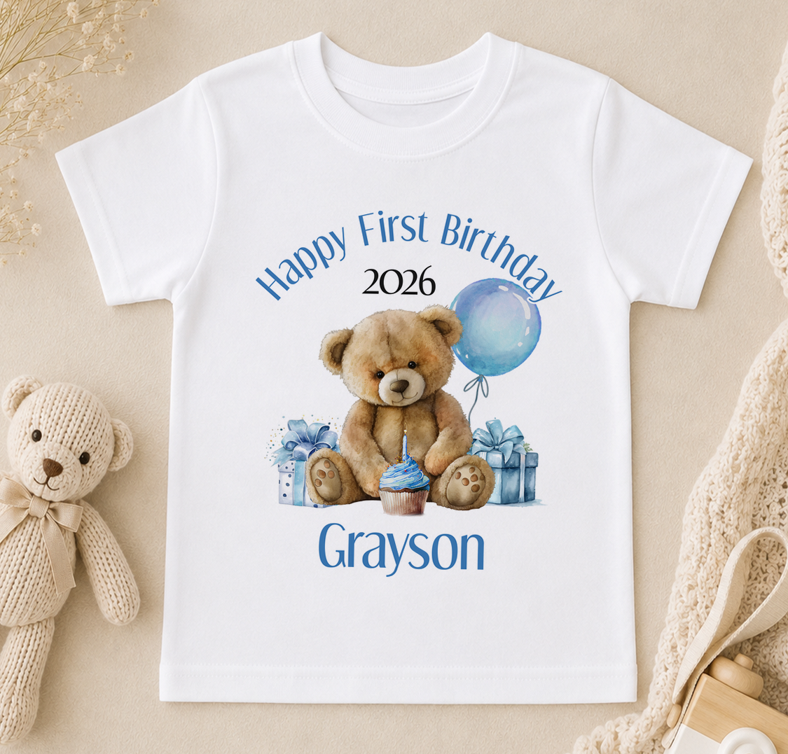 A white t-shirt with a printed design of a teddy bear and birthday decorations, along with the text "Happy First Birthday Grayson" printed on it.