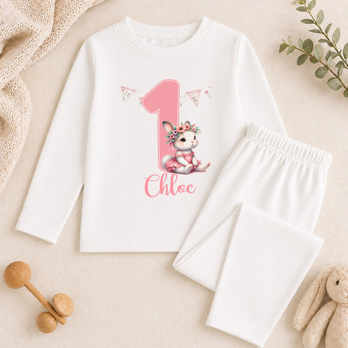 A white long-sleeved shirt with a pink bunny graphic and the name " Chloe" printed on it, along with a pair of white pajama pants. The shirt and pants are placed on a beige surface, and there are some stuffed animals and a plant in the background.