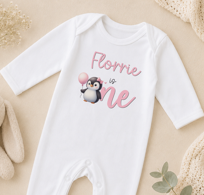 A white onesie with a penguin graphic and the text "Flornie is one" printed on it, along with a balloon.