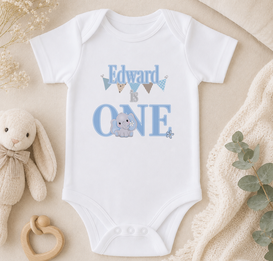 A white onesie with the text "Edward is one" printed on it, along with an image of a baby elephant. The onesie is placed on a beige surface, surrounded by a stuffed animal, a wooden toy, and some greenery.