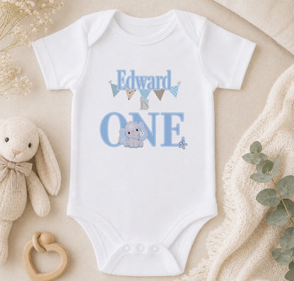 A white onesie with the text "Edward is one" printed on it, along with an image of a baby elephant. The onesie is placed on a beige surface, surrounded by a stuffed animal, a wooden toy, and some greenery.