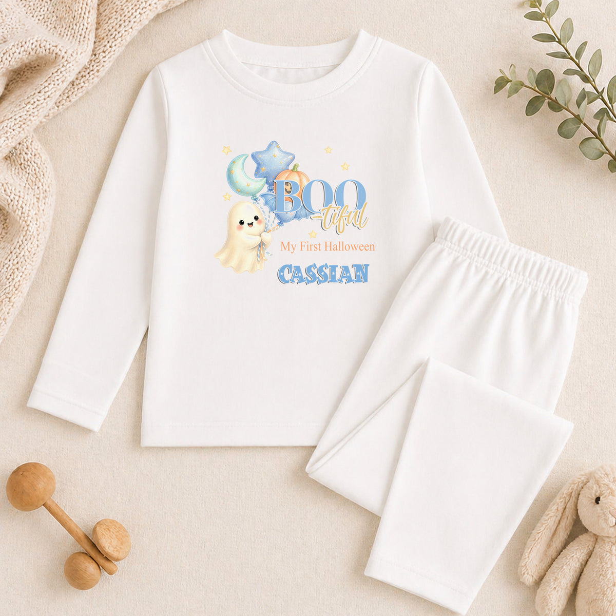 A white long-sleeved shirt with a Halloween-themed graphic and text, along with a pair of white pajama pants, and some stuffed animals on a beige background.