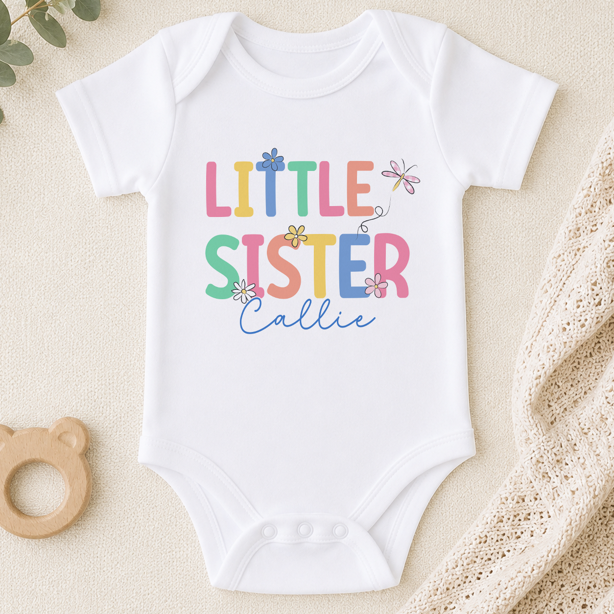 A white baby onesie with the text "Little Sister" printed on it, along with colorful flowers and a bear-shaped binky.
