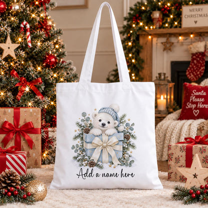 White cotton tote bag printed with a watercolour polar bear cub in a bobble hat peeping from a blue present, above a name.