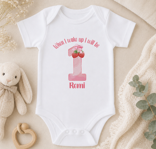 A white onesie with a pink number one and the text "When I wake up I will be" printed on it, along with the name "Rami" below the number. The onesie is placed on a beige blanket, and there is a stuffed animal and some greenery in the background.