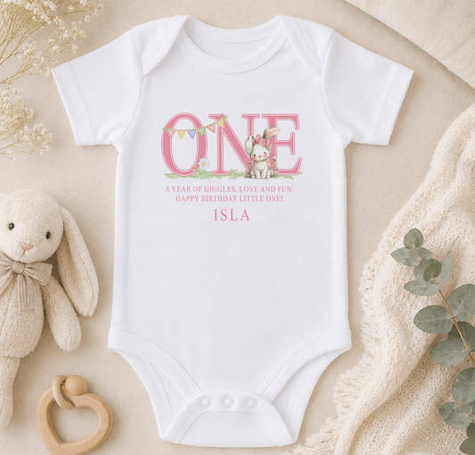 A white onesie with a pink bunny and the text "ONE" printed on it, along with a personalized message, is displayed on a bed with a stuffed bunny and a blanket.