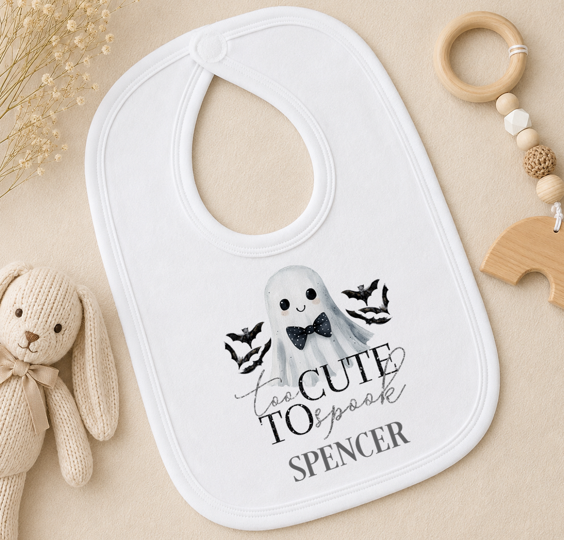 A white baby bib with a ghost design and the text "To Cute to Speak" printed on it, along with a wooden toy and a wooden bead ring.