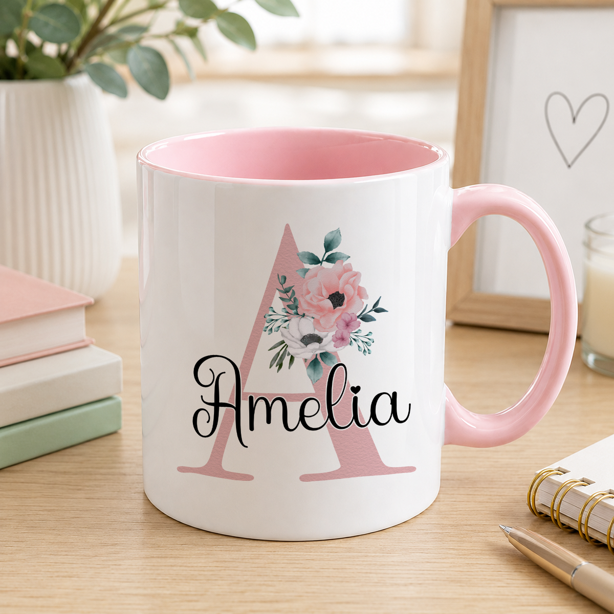 A pink and white mug with the name "Amelia" printed on it, sitting on a wooden surface next to some books and a plant.