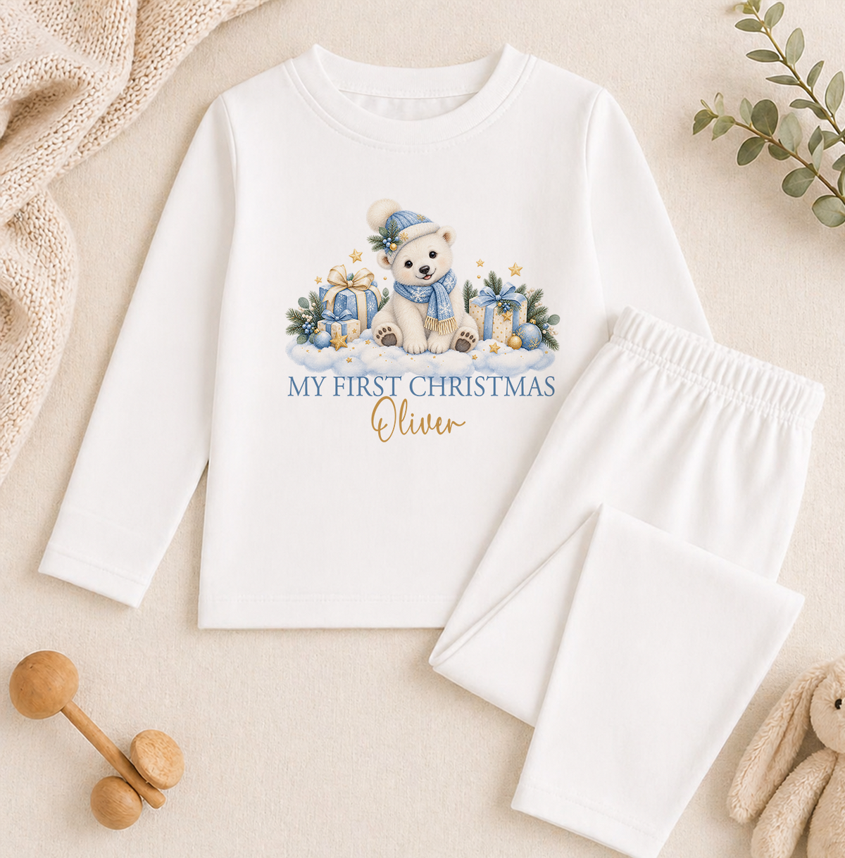 A white long-sleeved shirt with a graphic of a polar bear and Christmas presents, along with the text "My first Christmas Oliver" printed on it. The shirt is placed on a beige surface, and there are some wooden toys and a plant in the background.