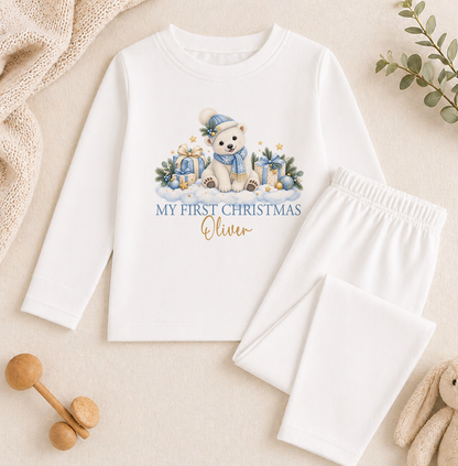 A white long-sleeved shirt with a graphic of a polar bear and Christmas presents, along with the text "My first Christmas Oliver" printed on it. The shirt is placed on a beige surface, and there are some wooden toys and a plant in the background.