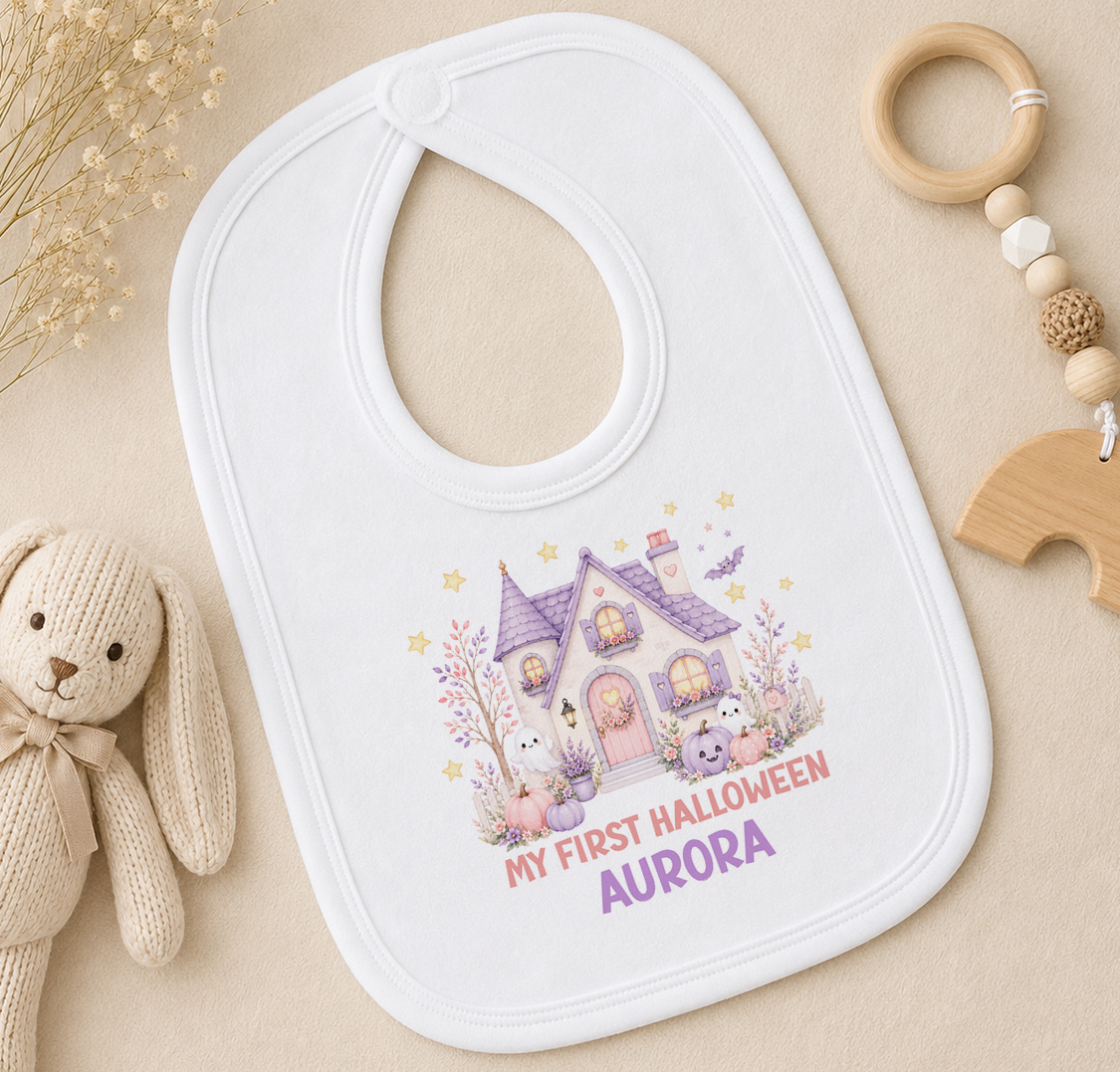 A white baby bib with a Halloween-themed design, featuring a house, bats, and other spooky elements, along with the text "My first Halloween Aurora".
