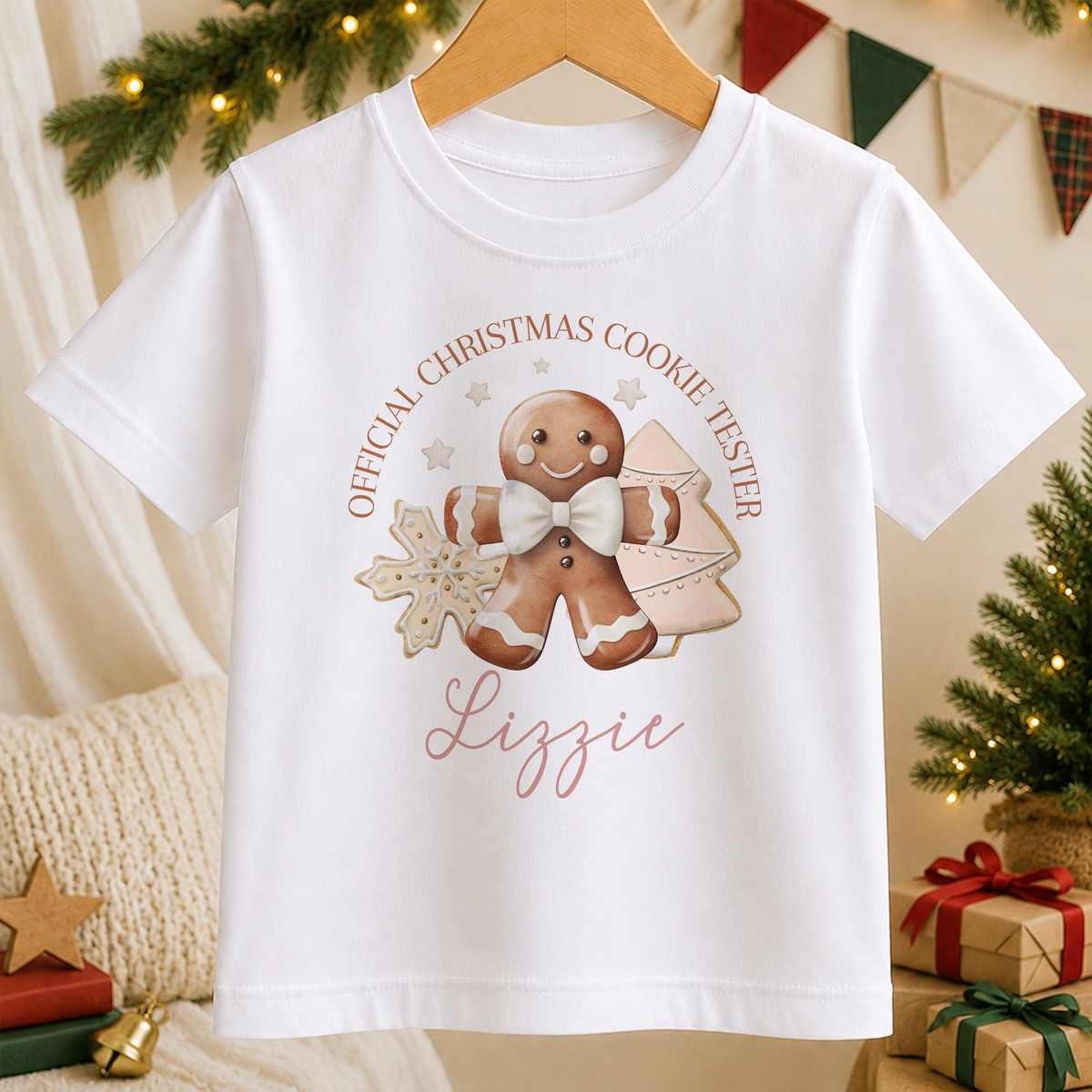 A white t-shirt with a Christmas-themed graphic design featuring a gingerbread man, snowflakes, and the text "Official Christmas Cookie Tester Lizzie".