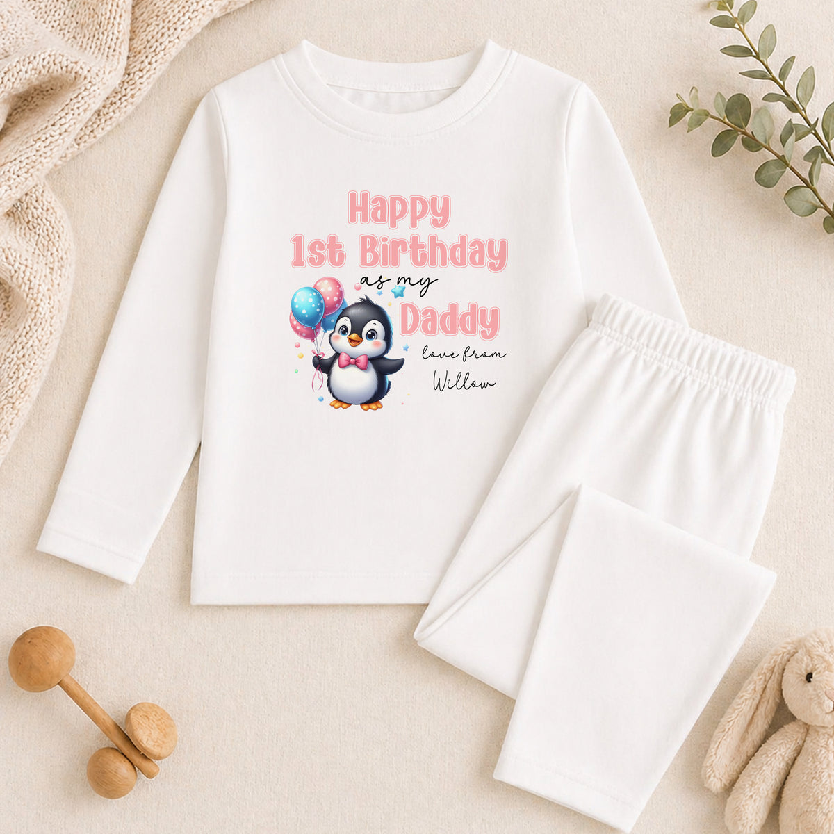 A white long-sleeved shirt with a penguin graphic and text, along with a pair of white pants, a stuffed animal, and some wooden toys, all arranged on a beige surface.