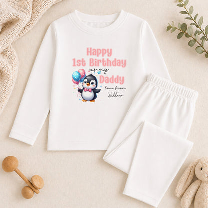 A white long-sleeved shirt with a penguin graphic and text, along with a pair of white pants, a stuffed animal, and some wooden toys, all arranged on a beige surface.