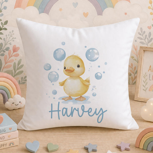 A white cushion printed with a yellow duckling among bubbles on a soft pink background, personalised with a child's name, photographed on a wooden surface among nursery toys and decorations.