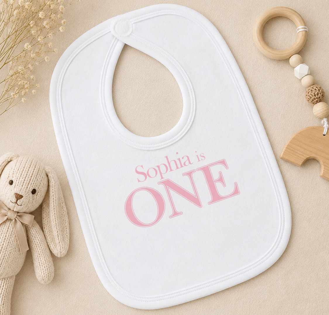 A white bib with the text "Sophia is ONE" printed on it, along with a wooden toy and a wooden bead ring.