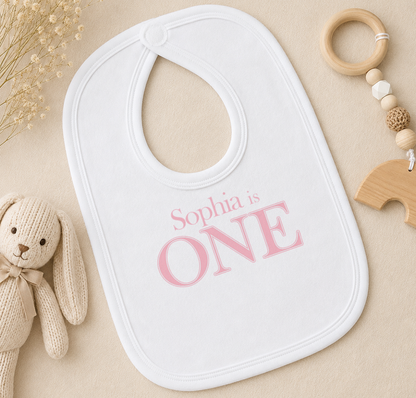 A white bib with the text "Sophia is ONE" printed on it, along with a wooden toy and a wooden bead ring.