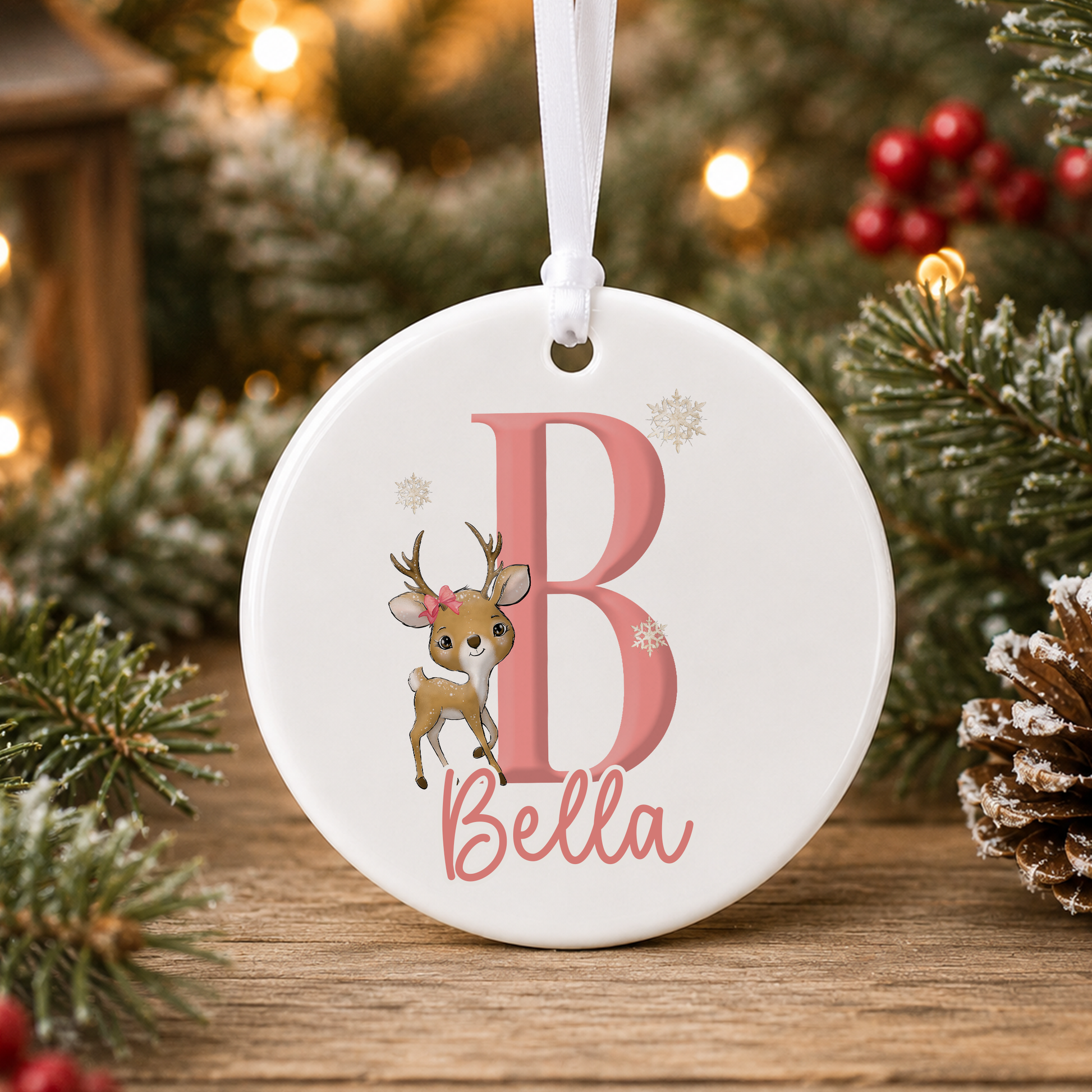 A white oval ornament with a pink letter "B" and a deer illustration, hanging on a Christmas tree.