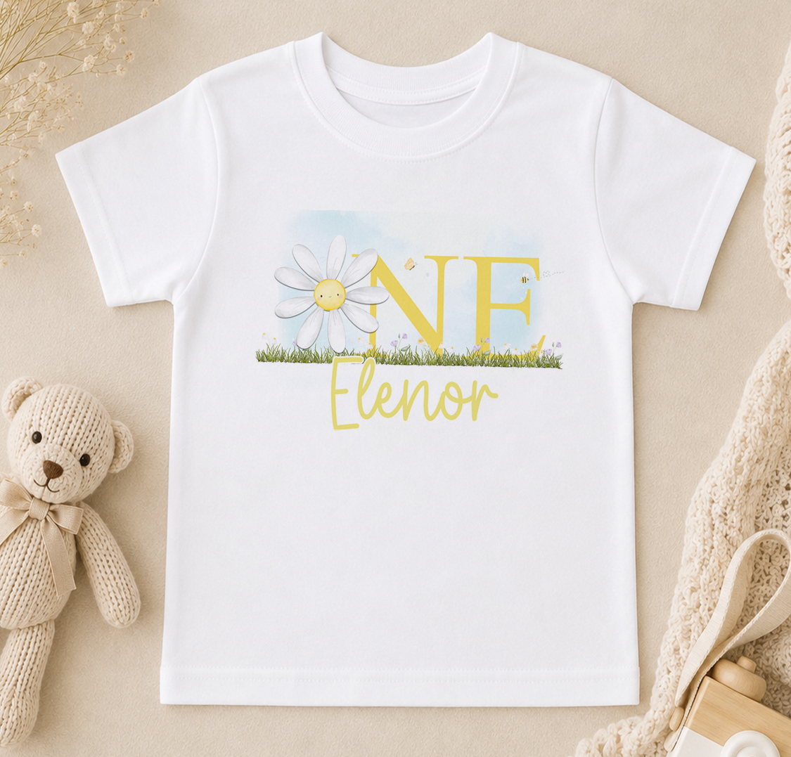 A white t-shirt with a yellow flower design and the word "Elenor" printed on it, along with a teddy bear and other baby items in the background.