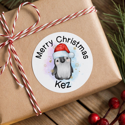 Round glossy Christmas sticker featuring a grey penguin wearing a red Santa hat, personalised with a custom name, shown on a wooden surface with pine and berries