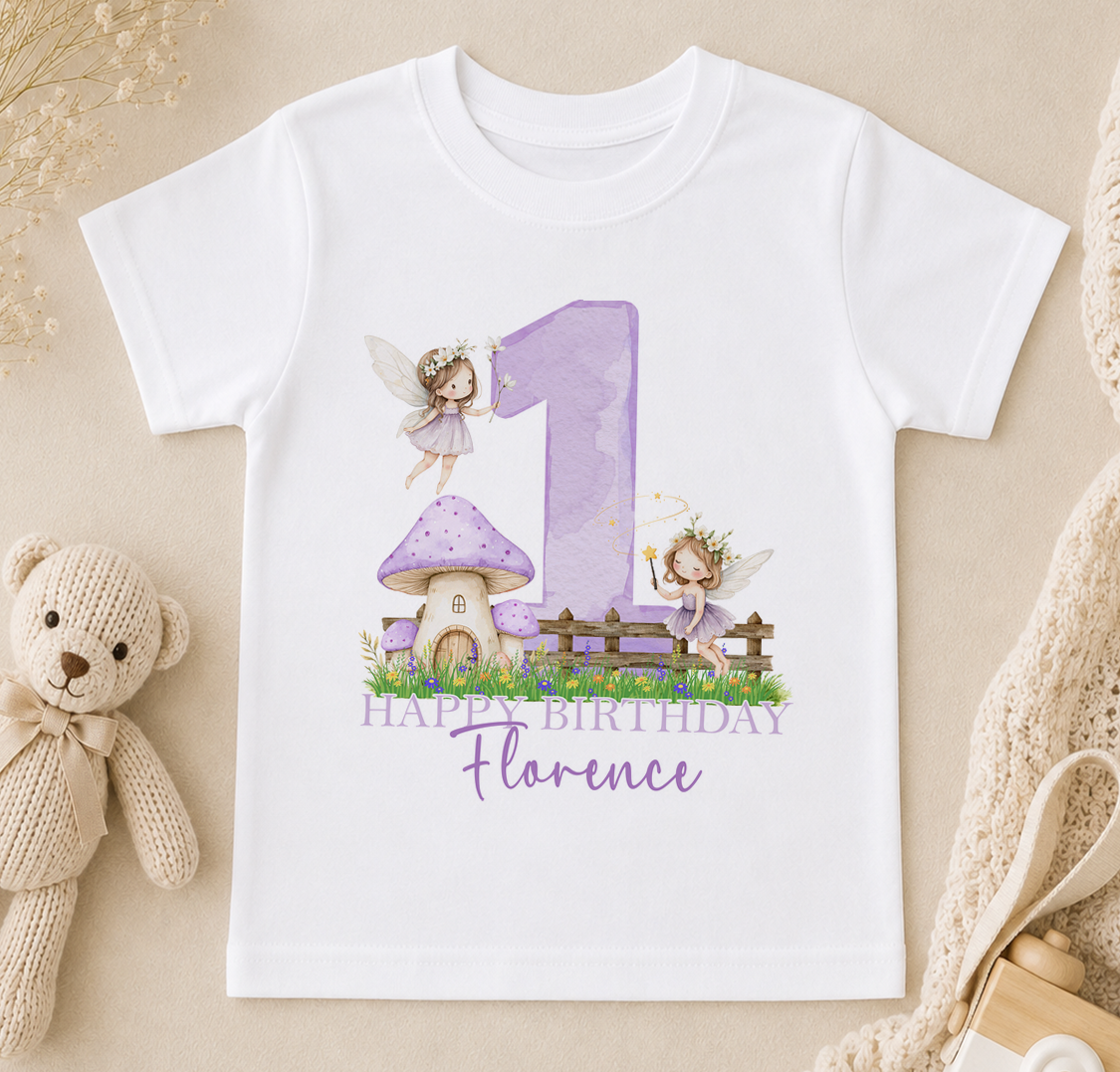 A white t-shirt with a purple number one and two-tailed fairies, along with a mushroom house and a "Happy Birthday Florence" message, is displayed on a beige surface.