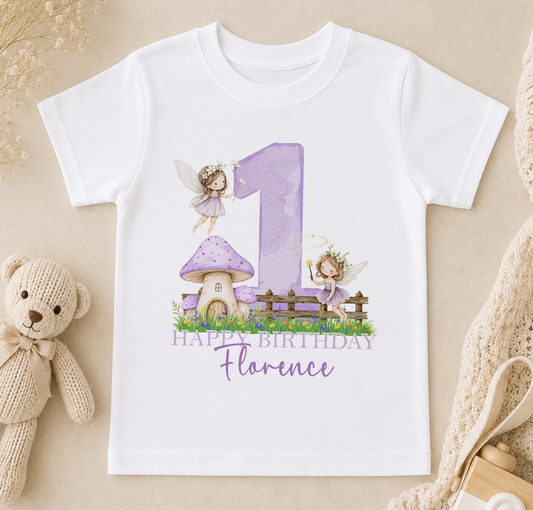 A white t-shirt with a purple number one and two-tailed fairies, along with a mushroom house and a "Happy Birthday Florence" message, is displayed on a beige surface.