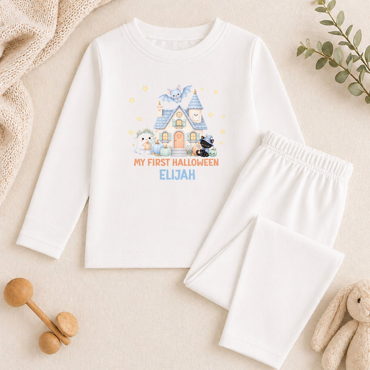 A white long-sleeved shirt with a Halloween-themed graphic and text, along with white pajama pants, a stuffed animal, and a wooden toy car, all arranged on a beige surface.