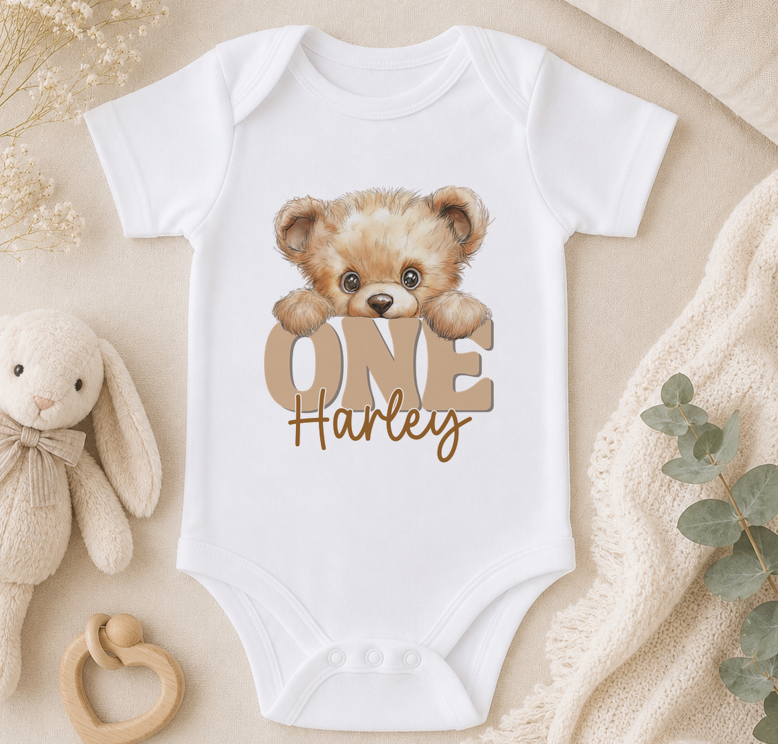 A white onesie with a teddy bear and the text "ONE Harley" printed on it, along with a wooden bib and a stuffed bunny toy.