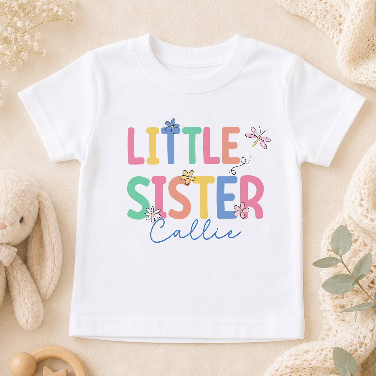 A white t-shirt with the text "LITTLE SISTER CALLIE" printed on it, surrounded by various baby-related items such as a stuffed animal, a blanket, and a toy.