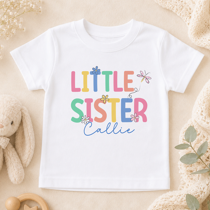 A white t-shirt with the text "LITTLE SISTER CALLIE" printed on it, surrounded by various baby-related items such as a stuffed animal, a blanket, and a toy.