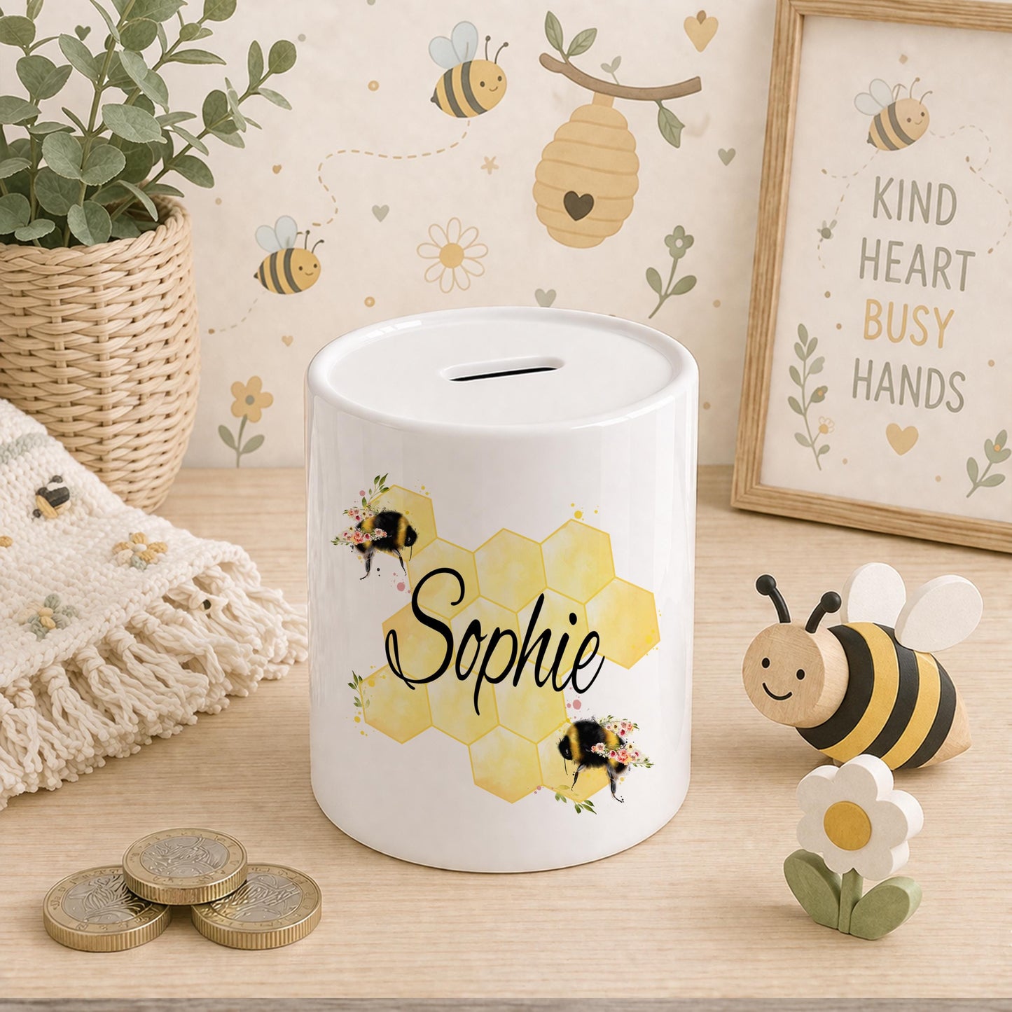 Ceramic bee money box with a honeycomb pattern and bumblebee illustrations, personalised name in black script, styled with a bee-print blanket, wooden bee toy and coins