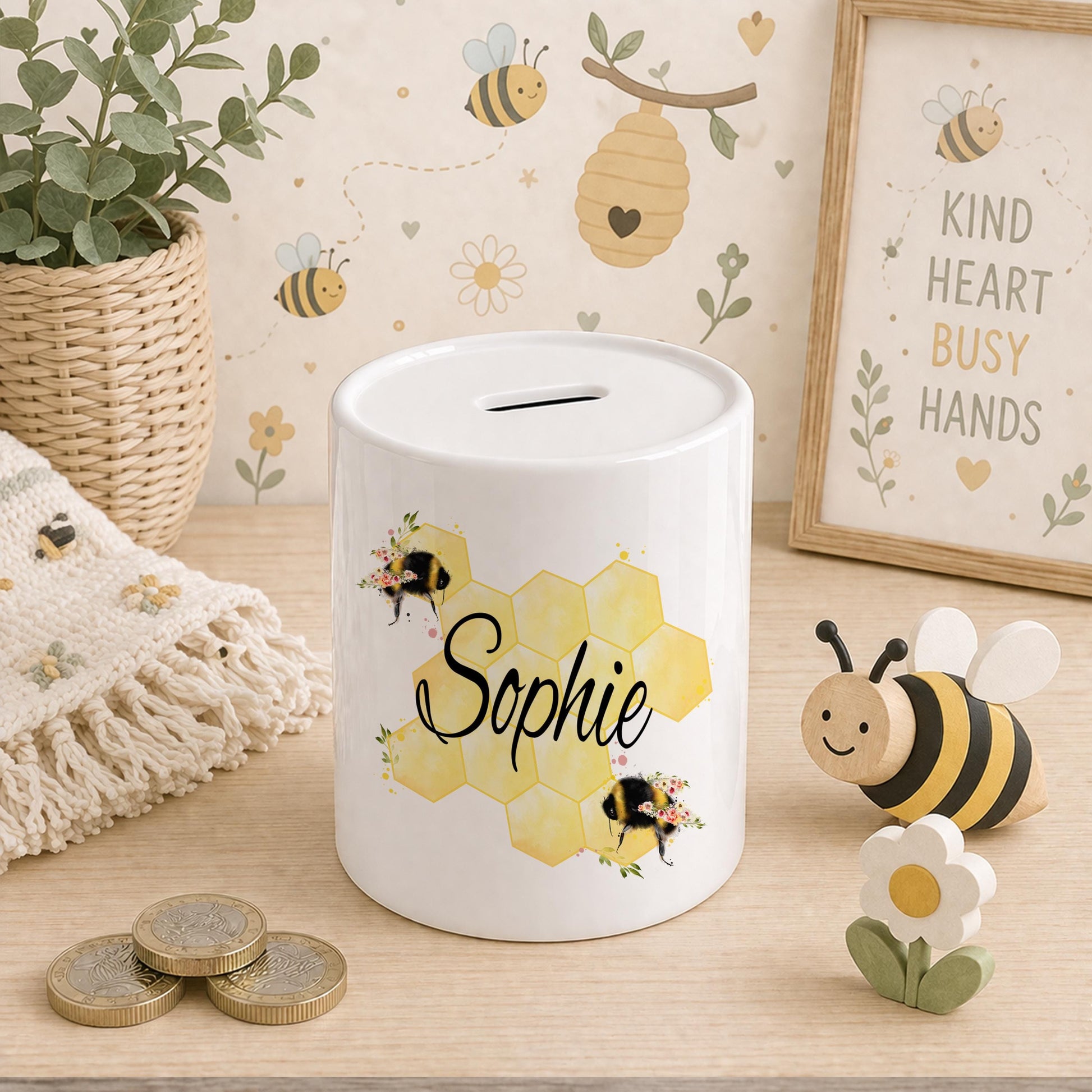 Ceramic bee money box with a honeycomb pattern and bumblebee illustrations, personalised name in black script, styled with a bee-print blanket, wooden bee toy and coins