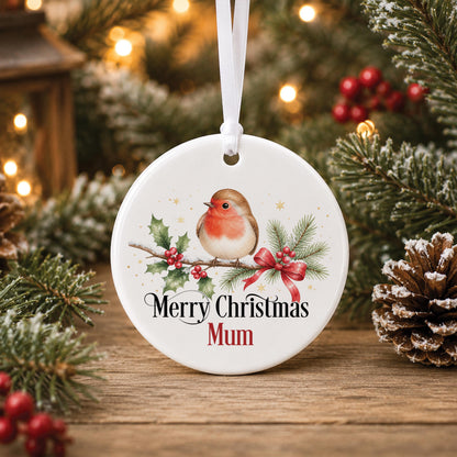 Personalised robin Christmas bauble, white ceramic ornament with a robin on a holly branch, printed with Merry Christmas Mum, hanging beside pine and pinecones