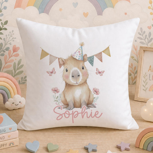 White cushion printed with a cartoon capybara wearing a party hat, surrounded by flowers, butterflies, a rainbow and a cloud, with the personalised name 'Sophie', styled on a wooden surface.