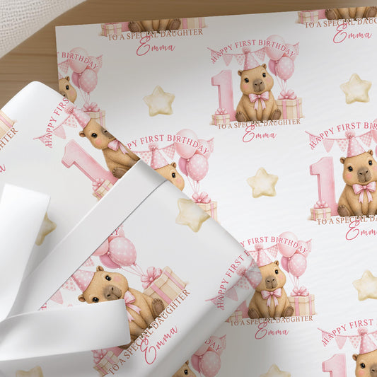 Close-up of personalised first birthday wrapping paper featuring a cute baby capybara, pink number one, balloons and presents on premium white wrapping paper.