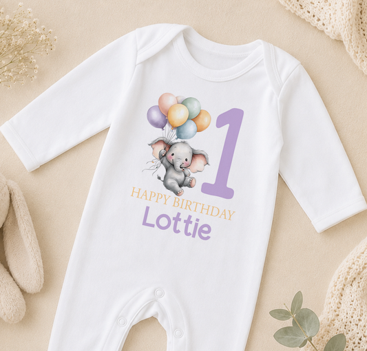 A white onesie with a cartoon elephant and balloons, along with the text "Happy Birthday Lottie" printed on it.