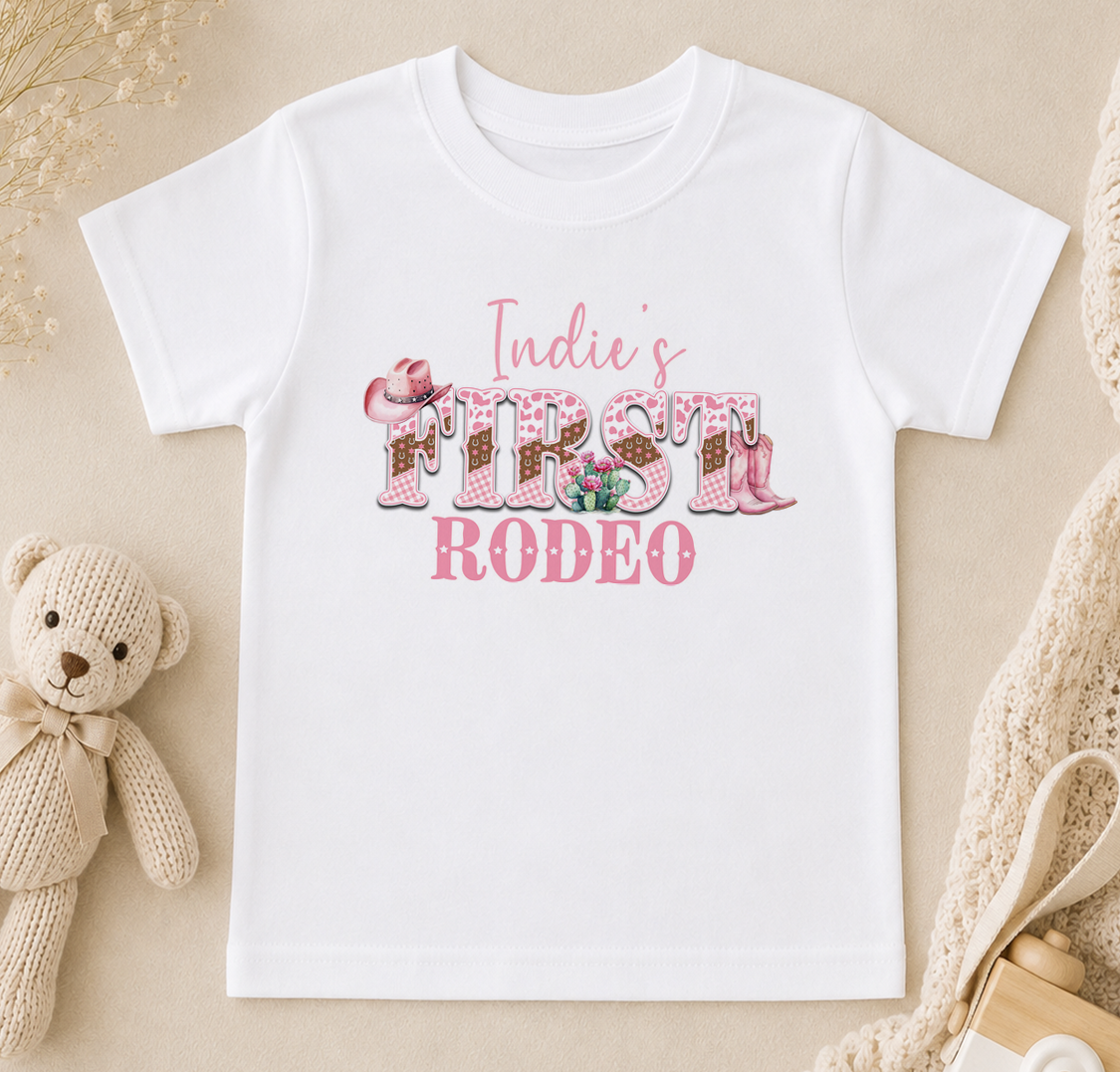 A white t-shirt with the text "Indie's FIRST RODEO" printed on it, along with various pink and white decorative elements such as flowers and cowboy boots.