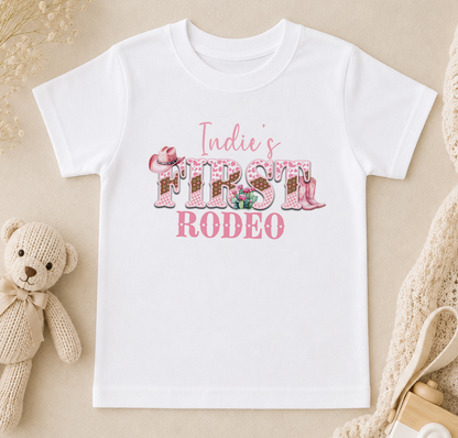 A white t-shirt with the text "Indie's FIRST RODEO" printed on it, along with various pink and white decorative elements such as flowers and cowboy boots.