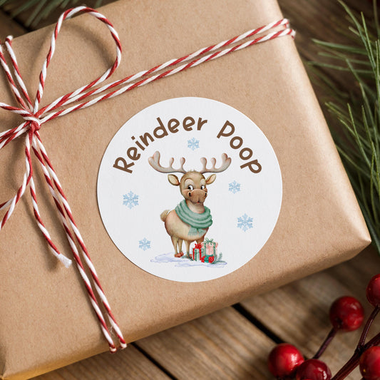 A brown gift box with a white label featuring a cartoon reindeer and the text "Reindeer Poop" on it, surrounded by Christmas decorations such as pine branches and berries.