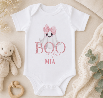 A white baby onesie with a ghost character and the text "BOOOUT MIIA" printed on it, along with a pink bow. The onesie is placed on a beige surface, surrounded by a stuffed animal, a baby rattle, and some greenery.