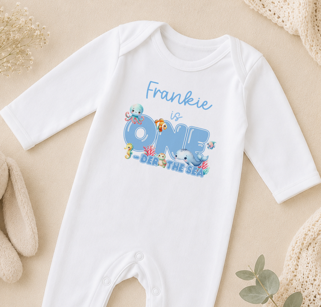 A white onesie with a personalized text and illustrations related to the ocean theme, specifically "Frankie is ONE DER THE SEA".