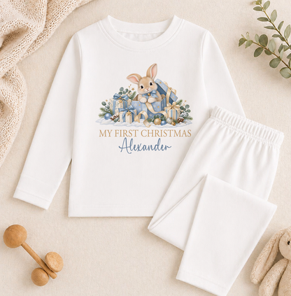 A white long-sleeved shirt with a bunny graphic and the text "My first Christmas Alexander" printed on it, along with a pair of white pajama pants. The shirt and pants are placed on a beige background, and there are some wooden toys and a plant visible in the image.