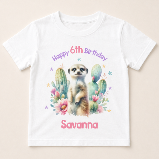 A white t-shirt with a printed design featuring a meerkat surrounded by cacti and flowers, along with the text "Happy 6th Birthday Savanna".
