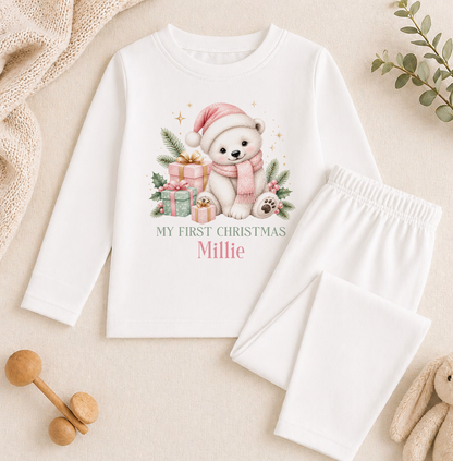 A white long-sleeved shirt with a Christmas-themed graphic of a polar bear wearing a Santa hat and holding presents, along with the text "My first Christmas Millie". The shirt is accompanied by white pajama pants and a small stuffed animal.